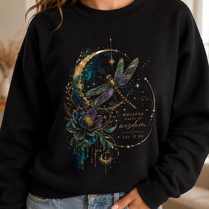 May include: A black long-sleeve sweatshirt featuring a graphic print of a dragonfly, a crescent moon, and a dark flower. The design includes gold accents, stars, and the text "WHISPER words of wisdom LET IT BE" in a mix of serif and script fonts.