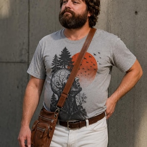 Puede incluir: Una camiseta gris jaspeado de manga corta con un estampado gráfico negro de una cabeza humana con ramas de árbol, un bosque y un gran sol rojo con pájaros volando. Se combina con pantalones blancos, un cinturón de cuero marrón y un bolso bandolera de cuero.