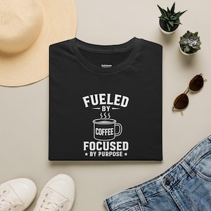 Peut inclure: Un t-shirt noir avec un motif blanc représentant une tasse de café et le texte "FUELED BY COFFEE FOCUSED BY PURPOSE" imprimé dans une police grasse sans empattement. Le design inclut des volutes de vapeur et deux petites étoiles en bas.