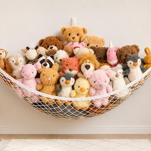 Op de afbeelding: Een witte speelgoedhangmat van gaas die aan een muur is bevestigd, gevuld met een verscheidenheid aan knuffels, waaronder beren, een leeuw, pinguïns, een varken en een konijn. Het net is bevestigd met drie witte haken voor opslag.