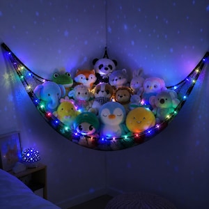 May include: A black mesh corner hammock filled with various plush animal toys, including a panda, elephant, penguin, and frog. The hammock is illuminated by a string of multicolored LED lights, casting a star-patterned projection on the walls.