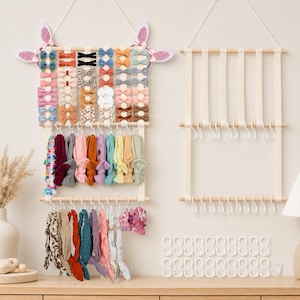 May include: Two wooden wall-mounted organisers with white hanging straps and hooks. The left organiser features glittery pink bunny ears and holds numerous small fabric bows, colourful headbands, and various scrunchies. The right organiser is empty.