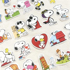 May include: A collection of die-cut vinyl stickers featuring the cartoon character Snoopy in various outfits and activities, including wearing a chef hat, pilot goggles, sunglasses, and holding items like a red heart, a book, and a sign reading Nice Swell Great Neat Splendid.