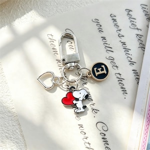 May include: A silver-toned metal keychain featuring a lobster clasp, a hollow heart charm, a circular charm with the letter E in gold on a black background, and a colorful enamel charm depicting a white dog holding a large red heart.