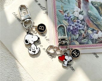 May include: Two silver-toned metal keychains featuring enamel charms of a white dog with black ears. One keychain includes a round black charm with the letter B, and the other features a red heart charm and a round black charm with the letter E.