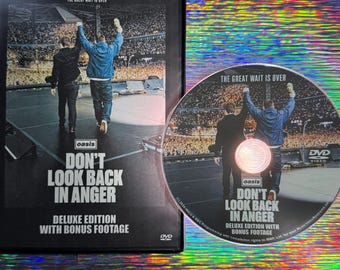 Puede incluir: Una caja de DVD negra y un disco circular a juego del concierto de Oasis Don't Look Back In Anger. La portada muestra a dos figuras en un escenario ante una gran multitud, con el texto The Great Wait Is Over y Deluxe Edition.