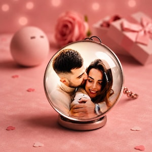 May include: A circular glass photo ornament featuring a printed image of a couple embracing. The ornament has a metallic rose gold frame and a matching circular base, designed to stand upright on a flat surface as a decorative display piece.