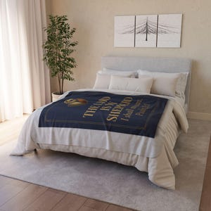 May include: A navy blue fleece blanket featuring a gold-coloured graphic of a shepherd's crook and the text "THE LORD IS MY SHEPHERD I shall not want. Psalm 23:1" draped over a bed with light grey bedding and white pillows.