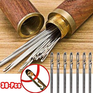 May include: A set of 30 silver-toned metal sewing needles with open-eye designs, stored in a cylindrical wooden case with a brass-colored threaded cap. The needles feature a side-opening eye for easy threading, displayed both inside the case and lined up.