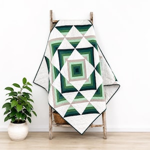 May include: A quilt featuring a geometric pattern of concentric squares and triangles in shades of dark green, forest green, sage green, and light beige, set against a white background. The fabric has a visible quilted texture throughout.