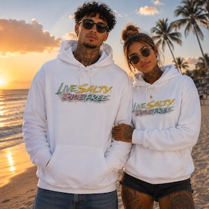 Könnte beinhalten: Zwei weiße Kapuzenpullover mit dem Schriftzug "LIVE SALTY RIDE FREE", der in einer stilisierten, mehrfarbigen Schrift auf der Brust gedruckt ist. Jeder Kapuzenpullover verfügt über eine Kängurutasche vorne, lange Ärmel und eine Kapuze.