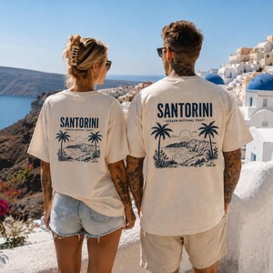 Könnte beinhalten: Zwei cremefarbene T-Shirts mit einer dunkelblauen Grafik auf der Rückseite. Das Design zeigt den Text "SANTORINI AEGEAN NATIONAL COAST EST 1500 BC" über einer Illustration von Palmen, einer Küstenlinie und einer aufgehenden Sonne.