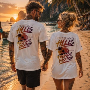 Könnte beinhalten: Ein weißes Kurzarm-T-Shirt mit einem Grafikdruck auf der Rückseite, der den Text Thailand Hills und Tropical Vacation zeigt. Das Design umfasst eine Sonnenuntergangsillustration mit Palmen in Orange-, Lila- und Gelbtönen.