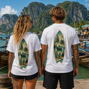 May include: Two white t-shirts featuring a surfboard-shaped graphic on the back. The graphic depicts a tropical scene with a long-tail boat, palm trees, a sun, and smaller surfboards in shades of green, blue, yellow, and brown against a white background.