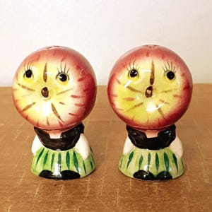 May include: A pair of ceramic salt and pepper shakers shaped like figures with round, apple-like heads featuring painted facial details, black tops, and green striped skirts. Each shaker has a smooth, glossy finish and a rounded base.
