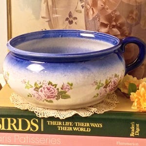 May include: A ceramic chamber pot with a blue rim and a floral rose pattern, resting on two stacked books titled Birds and Paris Patisseries. Beside it stands a brass candelabra with two yellow candles and a tall, light-colored floral vase.