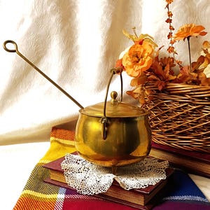 May include: A polished brass pot with a rounded body, a lid with a small knob, and a swinging handle. A long, thin brass rod with a circular loop at the end extends diagonally from the pot. The pot rests on three small feet atop a white lace doily.