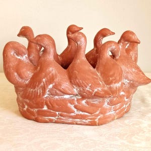 May include: A terracotta-colored ceramic planter featuring a circular arrangement of multiple duck figures facing outward. The base is textured to resemble a woven basket, and the entire piece has a matte finish with subtle white highlights.