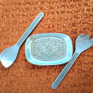 May include: A silver-toned metal serving set consisting of a small rectangular dish, a spoon, and a fork. The dish features a central spiral and star pattern, while the spoon and fork handles are decorated with geometric zigzag and spiral motifs.