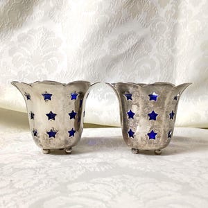 May include: A pair of silver-toned metal votive candle holders featuring a scalloped rim and a series of cut-out star shapes that reveal a deep blue interior lining. Each holder stands on three small, rounded feet, creating a stable base.