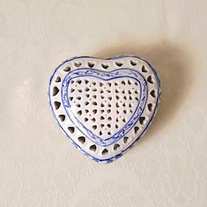 May include: A heart-shaped ceramic trinket box featuring intricate openwork cutouts in a lattice pattern. The white porcelain surface is decorated with delicate blue painted floral scrollwork along the edges and borders of the lid.