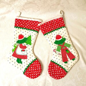 May include: Two quilted Christmas stockings featuring red and white patterned cuffs and toes. The center panels are white with small green leaf prints, each displaying a fabric applique of a child in red and green clothing holding a tree.