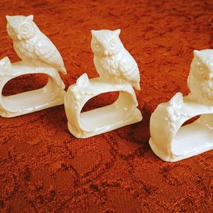 May include: A set of three off-white ceramic napkin rings, each featuring a detailed owl perched atop a branch. The owls have textured feathers and large eyes, while the base of each ring is shaped like a gnarled tree branch with small berries.