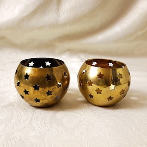 May include: Two spherical brass candle holders featuring a repeating cutout star pattern across the surface. The metal has a polished gold-toned finish with visible natural variations in the texture of the brass material.