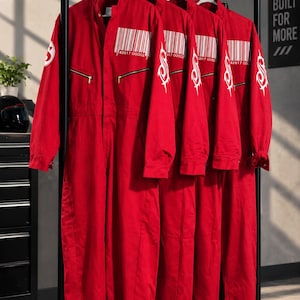 Puede incluir: Cuatro monos de color rojo brillante colgados en un perchero de metal negro. Cada mono presenta un gráfico de código de barras blanco con el número 42617 00002 7 en el pecho, un logo tribal blanco en la manga y bolsillos con cremallera dorada.