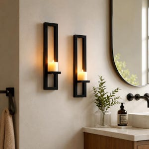 Peut inclure: Deux appliques murales rectangulaires en métal noir montées verticalement, chacune dotée d'une étagère centrale supportant une bougie pilier blanche allumée. Les appliques ont un cadre ouvert et sont fixées sur un mur clair au-dessus d'un meuble.