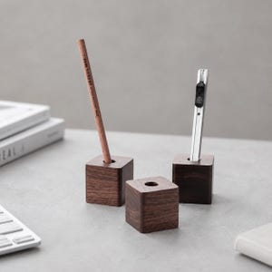 May include: Three dark brown wooden cube pen holders with a smooth finish. One holder contains a wooden pencil, another holds a silver metal utility knife, and the third cube remains empty. Each cube features a single circular hole on the top surface.