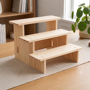 May include: A three-tiered wooden display stand featuring a light natural wood grain finish. The structure consists of three horizontal shelves arranged in a stepped formation, supported by vertical side panels with visible joinery details.