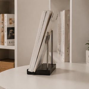 May include: A magazine rack featuring a rectangular black wooden base and two vertical silver metal rods that support a stack of white magazines. The magazines are positioned at an angle, held upright by the metal structure, displaying white covers.