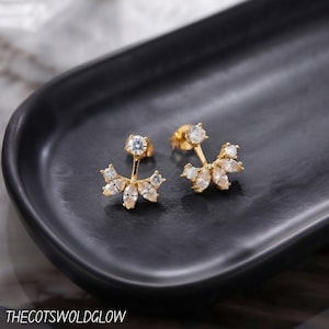 May include: A pair of gold-tone ear jacket earrings featuring a single round clear stone stud connected to a fan-shaped base adorned with marquise-cut clear stones. The text THECOTSWOLDGLOW is visible at the bottom of the image.