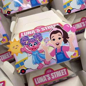 May include: A white cardboard favor box featuring a colorful graphic of a cartoon character with purple hair and a blue dress, alongside a cartoon person in a pink shirt and blue overalls. The text "LUNA'S STREET" is printed on a pink rectangular label.