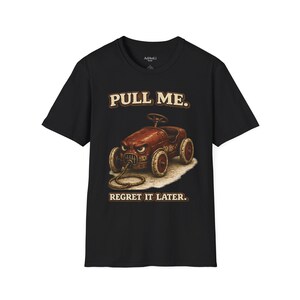 以下が含まれることがあります： 黒の半袖Tシャツ。怒った表情をした錆びた赤いおもちゃの車のグラフィックが中央にあり、車の前部にはロープが繋がれています。車の上部には「PULL ME.」、下部には「REGRET IT LATER.」という文字がヴィンテージ風のフォントでプリントされています。