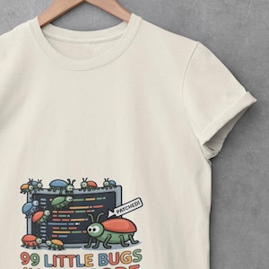 May include: A cream-colored t-shirt featuring a graphic of a computer screen with colorful bugs and the text 99 LITTLE BUGS IN THE CODE. One large bug in the foreground has a speech bubble that says PATCHED! The shirt is displayed on a wooden hanger.
