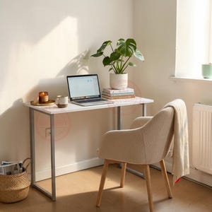 May include: A white rectangular desk with a silver metal frame, a laptop, a small potted monstera plant, a candle, and a mug. An off-white textured armchair with a draped blanket sits in front of the desk, and a woven basket is on the floor.