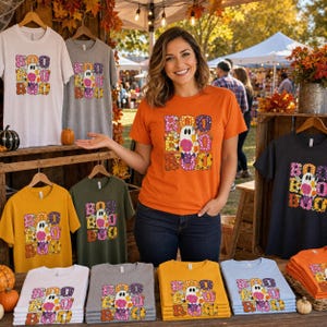 Puede incluir: Una colección de camisetas de manga corta en varios colores, incluyendo blanco, gris, naranja, verde oliva y azul claro. Cada camiseta presenta un estampado gráfico de un fantasma con el texto BOO en letras coloridas y estampadas.