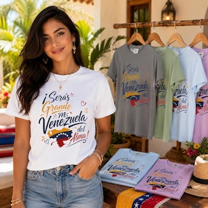 Puede incluir: Una camiseta blanca de manga corta con el texto "¡Serás Grande mi Venezuela del Alma!" en letra cursiva azul y dorada, acompañada por un gráfico de la bandera venezolana con la forma del mapa del país y pequeños corazones rojos.