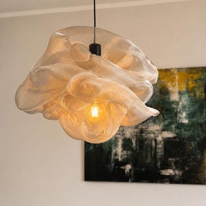 Könnte beinhalten: Eine Pendelleuchte mit einem halbtransparenten, cremeweißen, netzartigen Stoffschirm, der in geschichteten, organischen, wolkenartigen Falten geformt ist. Eine einzelne warm leuchtende Edison-Glühbirne ist in der Mitte sichtbar.