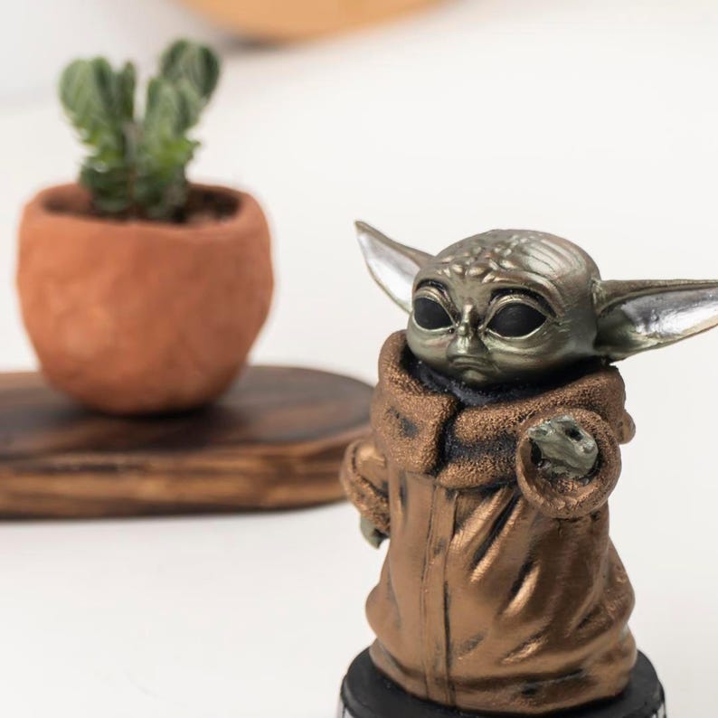 May include: A small, metallic bronze-colored figurine of a creature with large, pointed ears, wide dark eyes, and a textured robe. The figure stands on a circular black base, featuring a detailed head with visible skin folds and a prominent collar.