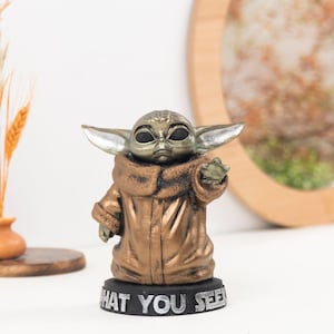 May include: A small, bronze-colored figurine of a creature with large pointed ears and dark eyes, wearing a textured robe. The figure stands on a black circular base with the silver-colored text "WHAT YOU SEE" embossed on the front edge.