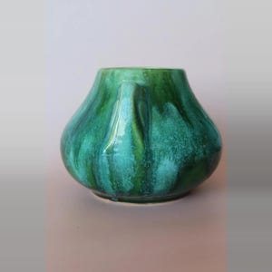 May include: A rounded ceramic vase featuring a glossy glaze with blended shades of emerald green, forest green, and teal. The vessel has a wide, bulbous body that tapers towards a narrow opening and includes a single vertical handle on the side.