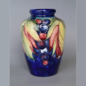 May include: A ceramic vase featuring a vibrant hand-painted design of dark blue berries and elongated leaves in shades of yellow, orange, and green. The base colour of the vase is a deep, rich blue, with a glossy finish and a rounded, bulbous shape.