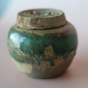 May include: A small, round ceramic jar with a lid, featuring a glossy, mottled emerald green glaze over a textured, earthy beige base. The surface displays a visible crackle pattern and uneven, rustic colour transitions across the rounded body.