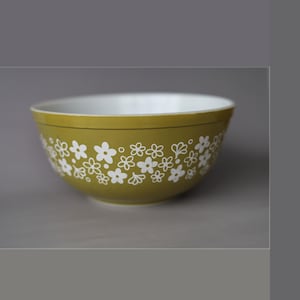 May include: A round mixing bowl with a smooth olive green exterior and a white interior. The exterior features a repeating pattern of small, white, five-petaled flowers and circular dots encircling the entire circumference of the bowl.