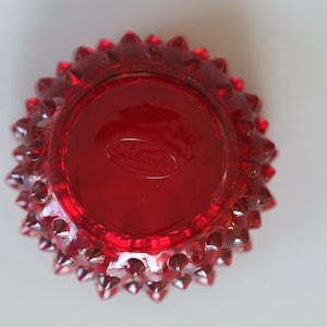 May include: A vibrant red glass bowl featuring a textured hobnail pattern on the exterior. The circular base is smooth and displays an embossed oval logo with the text Fenton, visible in the centre of the underside of the glass vessel.