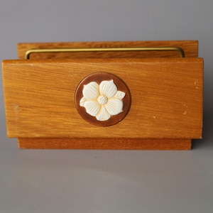 May include: A rectangular wooden napkin holder with a medium brown wood grain finish. The front panel features a circular inset with a white, five-petaled floral relief carving. A thin, gold-toned metal bar spans the top of the wooden structure.