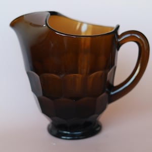 May include: A translucent amber-coloured glass pitcher featuring a repeating hexagonal honeycomb pattern across its body. The pitcher has a curved pouring spout, a sturdy handle on the side, and a circular base, all crafted from a single piece of glass.